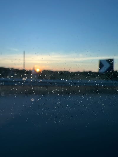 This image, captured in Cassano delle Murge, Italy, depicts an outdoor scene at either dawn or dusk, viewed through a transparent surface heavily covered with water droplets. The foreground is dominated by these sharply focused, spherical water droplets of various sizes on a window, creating a bokeh effect on the background elements.

The midground reveals a highly blurred landscape. A dark, undulating horizon line, likely composed of distant trees or hills, spans the width of the frame. A bright, intense orange-yellow orb, representing the sun, is positioned centrally on this horizon, partially obscured by the silhouetted terrain. To the left of the sun, a faint, tall, slender vertical structure, possibly a pole or mast, rises against the sky. To the right, a dark, blurred rectangular object with a prominent white chevron pointing left is visible, consistent with a road sign. Below the horizon line, a dark, elongated band suggests a road surface or barrier.

The background consists of the sky, which transitions from a light orange-yellow hue directly above the sun, indicative of twilight, to a clear, progressively deeper blue in the upper portion of the image. No distinct cloud formations are discernible. There are no people or explicit actions depicted. The scene suggests observation from a stationary or moving vehicle.