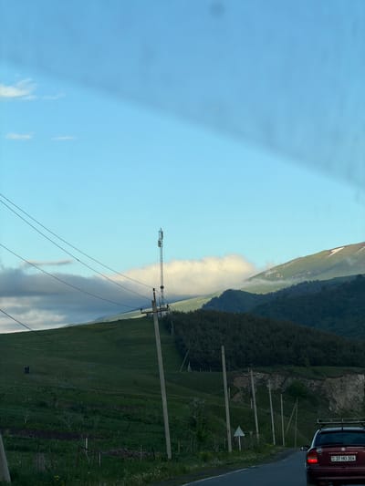 The image depicts a mountainous landscape in Fioletovo, Armenia, partially obscured by a low-lying cloud formation. The sky is blue with some light clouds. The land is green with vegetation, and power lines run along the roadside. A red vehicle is visible on the road in the upper right corner, with license plate number "37 HO 304." Tall, wooden utility poles stand along the right side of the image.