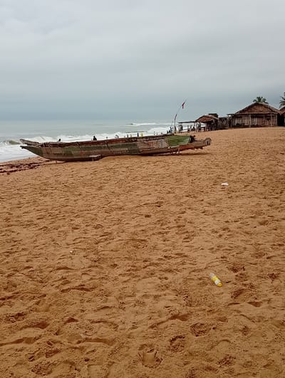 The image depicts a coastal scene in Gbaj, Nigeria. A large, weathered wooden boat rests on a sandy beach. The boat appears old, with visible wear and tear, and is positioned close to the shoreline where waves are breaking. In the background are several thatched-roof structures, likely beach huts or dwellings. The sky is overcast, suggesting a cloudy or potentially rainy day. Litter is scattered on the beach. People are visible near the boat and in the vicinity of the thatched-roof structures, but their activities are not clearly discernible.