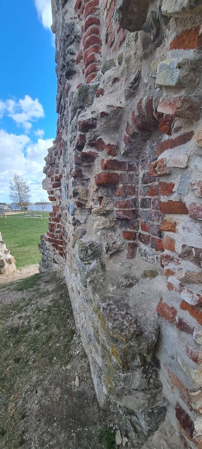 The image shows a brick and stone structure, likely a ruin, located in Salaspils, Latvia. The structure's construction suggests an old building or part of a fortification, featuring a mix of red bricks and grey stones held together with mortar. The sky is visible through an opening in the structure, showcasing a blue sky with scattered white clouds. The lower portion of the image reveals a grassy area, indicating the structure is situated in an open, possibly park-like setting. A tree and a fence can be seen in the background.