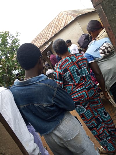 The image captures a gathering of people outside a building with a corrugated metal roof. The location is identified as Panyam, Nigeria. In the foreground, a person wearing a denim jacket is observed from behind. To their right, another individual sports a vibrant, patterned outfit consisting of a top and pants with geometric designs in red, blue, and black. Further in the background, several other individuals are visible, some wearing white shirts and others in darker clothing. A person in a light blue shirt with text that appears to read "MOTTO: SUCCESS" is also present. The environment suggests an outdoor, possibly informal, community event or meeting. The ground appears to be a dirt or sandy surface.