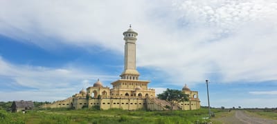 The image shows the Baiturrahman Mosque, also known as the Grand Mosque of Kruengmate, located in Kruengmate, Indonesia. The mosque is a prominent structure with a tall, slender minaret and several domes. The building is painted a pale yellow and is surrounded by lush green vegetation and a field of rice paddies. The sky is blue with scattered white clouds. In the foreground, to the left of the mosque, there is a small, dilapidated wooden structure with a thatched roof. To the right of the mosque, a dirt road leads into the distance, with a utility pole and wires present. There is a bird flying in the sky above and to the left of the mosque. The overall scene depicts a peaceful rural landscape with a significant religious building.
