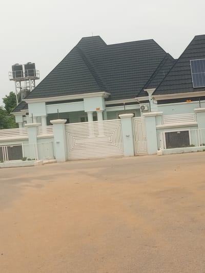 The image shows a light blue house with a dark gray tiled roof. A large white metal gate with a geometric design is in front of the house. The gate is flanked by white pillars. To the left of the gate is a white fence with metal grates. Behind the house, on the left, are two large black water tanks on a metal structure. A solar panel is visible on the roof of the house on the right. The house appears to be modern in style. The ground in front of the house is a dirt road. The sky is overcast, suggesting a cloudy day. The overall scene depicts a residential property. There is no visible text in the image. The location context is Baccure Kofare, Nigeria.