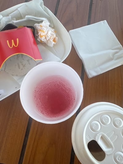 The image depicts a close-up, top-down view of a fast-food meal on a wooden surface. In the center is a white paper cup filled with a red beverage, condensation visible on the outside. To the upper left, a red and yellow McDonald's fry container is seen, with a crumpled napkin or wrapper next to it. To the right of the cup, a stack of white paper napkins is arranged neatly. In the lower right, a white plastic lid for a cup is visible, with embossed numbers and a central opening. The overall scene suggests a recently consumed meal. The location in Noyon, France is not discernible from the image.