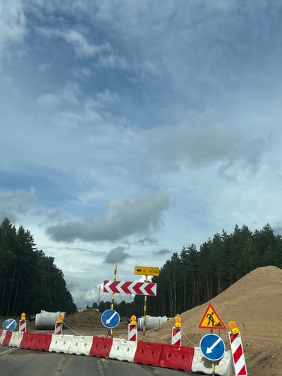 The image captures a section of road under construction, indicated by traffic barriers, signage, and a large pile of sand. The scene is located in the Плешчаніцкі сельскі Савет, Belarus, as suggested by the road signs. The road itself appears to be wet, possibly from recent rain.
On the left side of the road, a line of red and white plastic barriers marks the construction zone. Behind these barriers, large concrete pipes are visible, suggesting underground infrastructure work. A dark-colored vehicle is partially visible behind the pipes.
Several traffic signs are strategically placed to guide vehicles. A yellow sign with a black arrow pointing left reads "а - езд" which translates to "entrance" or "driveway". A large chevron sign with red and white arrows directs traffic to the right. Two blue circular signs with white arrows indicate a mandatory turn to the right. A yellow triangular sign depicts a person working with a shovel, signifying road work ahead. Additionally, two orange flashing lights are mounted on poles next to the barriers.
The background is dominated by a dense forest of pine trees lining the road on both sides. To the right, a steep mound of sand rises behind the construction area. The sky above is partly cloudy, with a mix of blue sky and scattered gray clouds. The overall impression is one of a road closure and ongoing construction project in a semi-rural or forested area.