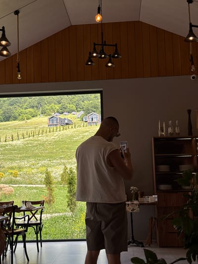 The image shows the interior of a restaurant in Dsegh, Armenia, with a large window offering a view of a lush green landscape. Inside, a man with a buzz cut, wearing a white sleeveless shirt and grey shorts, stands facing away from the camera, holding a mobile phone to take a photo of the scenery. He is positioned near a wooden cabinet filled with dishes and decorative items, including bottles and a peppermill. In the foreground, several wooden chairs surround a table set for dining. The restaurant features a high ceiling with visible wooden beams and industrial-style pendant lights. Outside the window, a rolling green hillside is dotted with scattered modern houses and solar panels, with trees and wildflowers creating a picturesque backdrop. The overall scene conveys a sense of modern rustic ambiance and a connection with the natural surroundings.