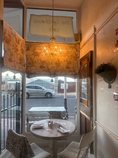 The image captures the inviting interior of what appears to be a cafe or restaurant, looking out onto a street scene through a large window. The perspective is from inside the establishment, oriented vertically in the provided image but described here as if upright.

**Subjects:**
The interior features a round, light-colored wooden dining table set for one, with a "RESERVED" sign, a wine glass, and cutlery on a napkin. An upholstered chair with a patterned fabric is visible to the right of the table. A multi-arm chandelier with exposed bulbs hangs from the ceiling, casting a warm glow, and patterned curtains or blinds with a toile-like design partially cover the windows. Above the window frame, a decorative wall hanging in the shape of a funnel holds artificial greenery. A speaker is mounted on the wall above another window/door frame to the left. The ceiling is plain white, with some molding detail.
Through the window, a street is visible, with a silver SUV parked along the curb. Parts of a decorative metal gate or fence are seen in the foreground outside. Further down the street, brick buildings and a lamppost are discernible. No people are visible inside or outside.

**Activity or event:**
The scene depicts a set dining table, suggesting the cafe is either open for business, preparing for guests, or has just closed. No active event is taking place; it's a still life moment.

**Notable details:**
The time of day appears to be daytime, with natural light entering through the large windows, though the interior lighting adds warmth. The atmosphere inside seems cozy and traditional, with the patterned fabrics and chandelier. The "RESERVED" sign on the table indicates the anticipation of a diner. A red sign with white text, possibly "TO" and an arrow, is visible on the upper wall, suggesting a directional indicator. The interior appears well-maintained, with a rustic yet elegant decor.

**Visible text:**
On the table, a small card clearly reads "RESERVED".
On the glass window, there is white text written in reverse, which appears to say "Cafe" followed by other, less decipherable words, possibly a French name or description.
On a red sign at the top of the image, the letters "TO" and an arrow are visible.