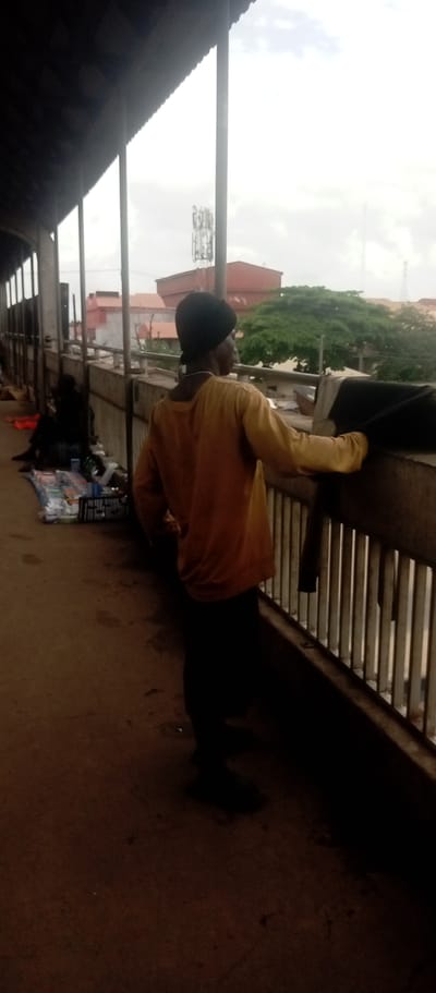 The image depicts a person standing on an elevated walkway, likely a pedestrian bridge, in Idemili, Nigeria. The person is wearing dark pants, a light orange long-sleeved shirt, and a dark head covering. They are leaning on the railing of the bridge, looking out at the cityscape. Other people are present nearby. Merchandise is set up next to the walkway, indicating possible informal trading. The environment includes buildings, trees, and communication towers in the background, under an overcast sky.