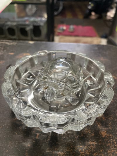 The image shows a clear glass ashtray with a sculpted turtle inside, placed on a dark, textured surface. The ashtray has a fluted rim and internal starburst patterns. The background is blurred, showing shelves with various items and a hint of red fabric. The lighting suggests an indoor setting, possibly a shop or display area. This scene is located in Lambha, India.