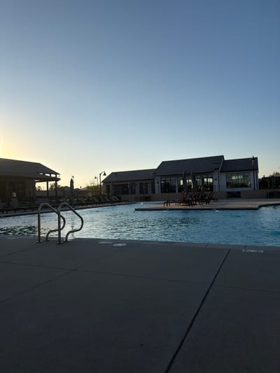 The image depicts a poolside scene at dusk in Santa Clarita, USA. The foreground shows the edge of a swimming pool with a set of chrome handrails leading into the water. The pool surface is rippled, reflecting the soft light of the setting sun. Adjacent to the pool is a concrete patio.
In the mid-ground, a building with large windows and a dark roof is visible, likely a clubhouse or community center. Several lounge chairs and umbrellas are arranged on the patio near the building, suggesting a recreational area. The sky above is a gradient of pale blue, transitioning to a lighter hue near the horizon, indicative of either sunrise or sunset.
Further back, more buildings and some landscaping are discernible against the twilight sky. The overall atmosphere is tranquil and serene, hinting at the end of a warm day. No people are visible in the image. A depth marker of "3 1/2" is visible on the pool's edge.