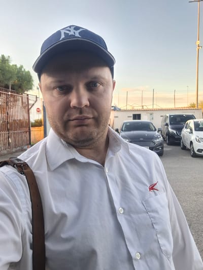 A man wearing a white collared shirt, a brown leather belt, and a dark blue baseball cap with a white "NY" logo is taking a selfie. He appears to be standing in a parking lot in the city of Quart de Poblet, Spain. There are several cars parked behind him. A chain-link fence and a building can be seen in the background. The sky is overcast.