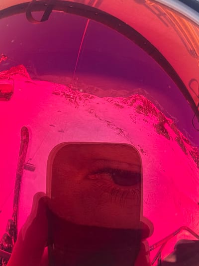 The image displays a first-person perspective taken from inside a ski lift, likely a gondola or bubble chairlift, with a curved structural frame visible at the top. The view outwards is through a strongly tinted, reddish-pink transparent surface, which dyes the entire external landscape and sky in corresponding hues. The scene depicts a snow-covered mountainous environment, indicative of a ski resort in Klösterle, Austria. Numerous ski lift cables and several individual chairlifts or cabins are visible traversing the slopes and ascending towards distant mountain peaks. The sky, viewed through the tint, appears deep purple. In the lower-center foreground, a distinct reflection of a human right eye, complete with visible eyelashes and iris, is present within a rectangular, light-edged object, consistent with a smartphone screen. A portion of a hand, presumably holding this object, is visible below the eye. To the left side of the frame, a segment of ski equipment, appearing as a ski or snowboard edge, is partially visible in an upright position. No specific individuals or their actions are discernable on the slopes, though the infrastructure indicates an active ski area.