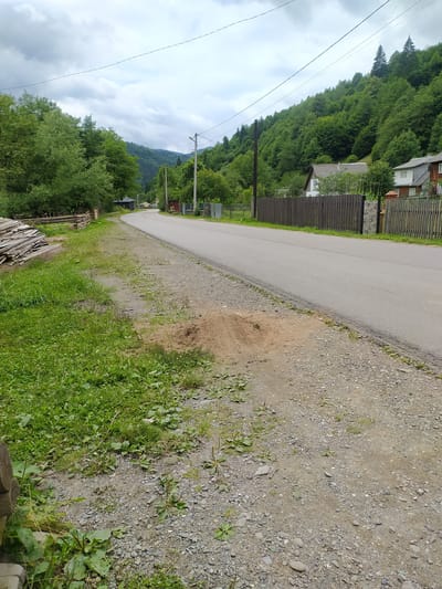 A paved road curves through a village in Breboia, Ukraine, bordered by lush green trees and a wooded hillside. On the left, a pile of weathered wooden planks lies near a rustic wooden fence. The road is flanked by a gravel shoulder with patches of grass and a small mound of dirt. Houses with fences are visible on the right side of the road, nestled among the trees. Overhead, a cloudy sky is crisscrossed by power lines.