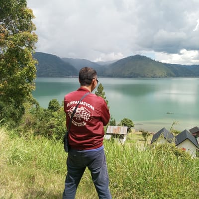 The image shows a person standing on a grassy hill overlooking a large lake surrounded by mountains. The person is wearing a maroon jacket with white writing on the back, dark pants, and a dark bag slung over their shoulder. They are facing away from the camera, looking out at the scenic view. The lake is a bright turquoise color, and the mountains in the background are covered in green vegetation. There are a few small buildings with dark roofs visible near the shore of the lake. The sky is cloudy. The location is identified as the city of Kalalengkio, Indonesia, which suggests this is a view of one of the many beautiful lakes in that region of Indonesia.