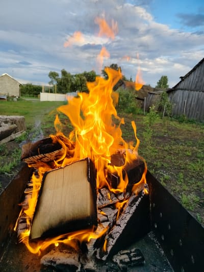 A fire burns brightly in a metal brazier, with large, yellow-orange flames reaching upwards. Several pieces of wood are visible, some with charred edges, nestled within the brazier. The brazier is placed outdoors in a grassy area with scattered dirt. In the background, there are several rural structures, including a weathered wooden building with a sloped roof and a smaller, lighter-colored building with a metal roof. Trees and green foliage are also visible, suggesting a natural or semi-rural setting. The sky above is cloudy, with patches of blue peeking through, indicating it might be late afternoon or early evening. The overall scene evokes a sense of warmth and an outdoor gathering or activity. There is no visible text in the image.