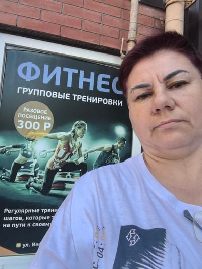 This is a medium close-up selfie of a middle-aged woman with short, dark red hair and a piercing above her left eyebrow. She is wearing a white t-shirt and has a somewhat neutral expression. Behind her is a large poster advertising a fitness club. The poster features the word "ФИТНЕС" (FITNESS) in large blue letters, followed by "ГРУППОВЫЕ ТРЕНИРОВКИ" (GROUP WORKOUTS). Below that, a circular graphic highlights a "РАЗОВОЕ ПОСЕЩЕНИЕ" (SINGLE VISIT) for "300 Р" (300 Rubles). The lower part of the poster shows images of people engaged in fitness activities, possibly on step platforms. The background brick wall and the metal pipe suggest an outdoor setting, likely in front of the fitness establishment. The poster also contains text in Russian, including "Регулярные трени шагов, которые т на пути к своему" and an address starting with "ул. Вен". The lighting suggests it is daytime.