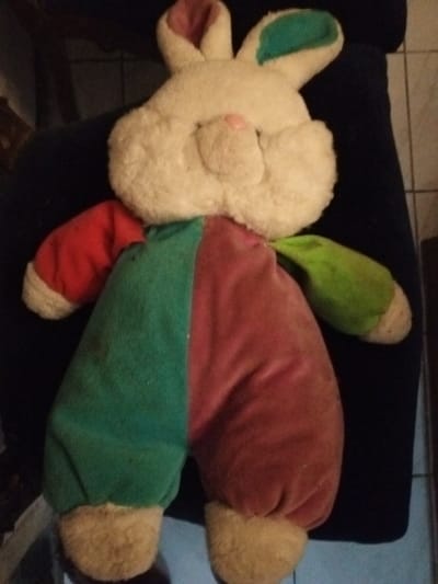A plush bunny doll with a cream-colored head and a pink nose sits against a dark background. The bunny's ears are tipped with green and pink. Its body is divided vertically into green and red sections. The bunny has red and green sleeves. Its feet are the same color as its head. The plush toy has a simplistic design.