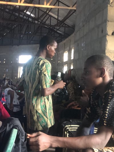 Two dark-skinned individuals are in close proximity, one wearing a green and yellow patterned shirt and the other wearing a black shirt with small gold designs and Adidas pants. They are indoors, appearing to be in a structure with a metal frame and what seems to be a cement ceiling. Based on the location provided, this is in Nteje, Nigeria.