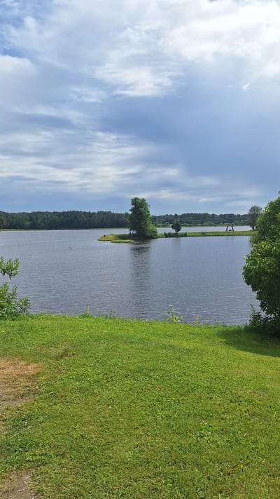 The image captures a serene waterscape in Stukmaņi, Latvia. A wide body of dark, rippling water dominates the center of the frame, bordered by lush green grass in the foreground. Across the water, a dense forest forms a dark green horizon line under a sky filled with dramatic clouds. A small, tree-covered island juts out into the water on the left side of the frame, with a solitary, tall tree standing prominently on it. Further in the distance, a swing set is visible on the opposite shore. The overall atmosphere is one of natural tranquility.
