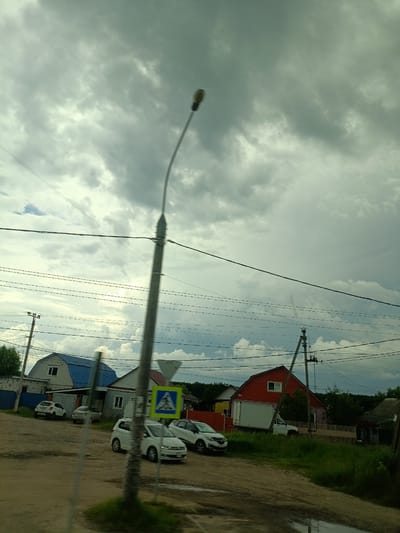 The image depicts a street scene in Saratovskaya, Russia, under a cloudy sky. A tall, grey street lamp with a curved arm is prominently positioned in the foreground, slightly to the right of the center. Multiple power lines crisscross the sky above a rural or suburban area.

On the left side of the image, a light-colored, two-story house with a bright blue roof is visible. Several cars are parked in front of it, including a white hatchback and a silver sedan. A pedestrian crossing sign, featuring a blue square with a white triangle and a pedestrian silhouette, is situated near the cars.

Further to the right, a red, two-story house with a white truck parked in front of it is seen. The truck appears to be a box truck. The surrounding vegetation consists of grass and trees, suggesting a semi-rural environment. The road surface appears to be dirt or gravel, with some wet patches indicating recent rain. The overall impression is of a typical day in a residential area, with overcast weather. There are no people clearly visible in the image.