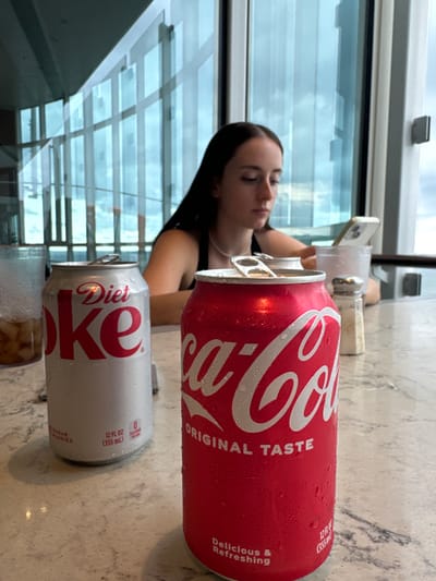This image depicts an indoor scene in Falmouth, Jamaica, likely a restaurant or lounge. The foreground features a light-colored, possibly granite, table surface. On this table, two aluminum beverage cans are prominent: a white "Diet Coke" can (12 fl oz / 355 mL) with condensation droplets and a closed tab, positioned to the left; and a red "Coca-Cola Original Taste" can (12 fl oz / 355 mL) with condensation droplets and an open tab, positioned to the right. A partially visible clear glass containing ice and a brown liquid is to the far left. A silver-topped salt shaker is located to the right of the red can.

In the midground, a young adult female with long, dark hair, wearing a dark top and a white choker necklace, is seated. She is observed looking down, engaged with a white smartphone that she holds in both hands and rests on a clear glass or cup. The background consists of large, multi-pane glass windows that reveal an overcast sky or a distant body of water, along with reflections of interior structural elements.