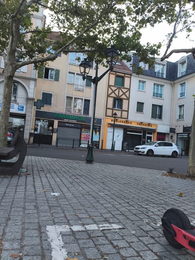 The image shows a street scene in Carrières-sous-Poissy, France, with buildings lining the street. In the foreground, a paved sidewalk is visible, with fallen leaves scattered on the ground. A red electric scooter lies on its side on the pavement. A dark-colored bench with an unusual design is also present on the sidewalk. Across the street, several storefronts are visible, including a "PHARMACIE SAINT-EXUPERY" and a "BOULANGERIE PATISSERIE". A white car is parked in front of the bakery. Trees with green leaves are overhead, and a lamppost stands on the sidewalk. The buildings appear to be multi-story residential or commercial properties with windows and shutters. The sky is overcast.