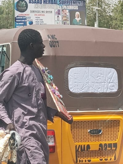 An individual, identified as a dark-skinned male, is depicted in profile, facing right, within the city of Hotoro, Nigeria. He wears a light purple long-sleeved garment and a dark cord necklace with an earbud-like attachment. He holds a flat, rectangular display board, adorned with multiple colorful, shiny items, possibly small accessories or jewelry, which he presents for sale. In his left hand, he carries a plastic bag containing white, textured items, possibly food products. The man's expression is neutral, and his gaze is directed forward.
Behind the individual is the rear of a yellow and brown motorized tricycle, commonly known as a Keke Napep or Auto Rickshaw. The brown fabric canopy of the vehicle displays the characters "KMC 0877" in black. A small, rectangular, translucent window with crumpled white material inside is visible on the rear canopy. The yellow lower section of the vehicle features a mesh grill with a silver "BE" emblem in the center. Below the grill, "KMC 0877" is also written in black, with additional, partially obscured characters below it. A red taillight is visible on the left side of the vehicle's rear.
Above the vehicle, a large billboard with a white background and blue text advertises "ASAAD HERBAL SUPPLEMENT AND ADULT STORE LTD & SPA SERVICES." Below this, a section titled "SERVICES WE OFFER" lists "SUPPLEMENT," "SLIMMING SUPPLEMENT," "WEIGHT GAIN SUPPLEMENT," and "FAT BURNER" on the left, and "SPA SERVICES," "FACIAL," "PEDI-CURE," "MANI-CURE," and "BODY MASSAGE" on the right. Visuals on the billboard include product images and a smiling woman wearing a headscarf. The scene is outdoors, under natural light, with discernible shadows.