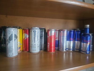 The image shows a wooden shelf with multiple cans of energy drinks. The shelf is filled with an assortment of brands and colors, including Red Bull, Monster, and other unspecified brands. The cans are arranged in rows, with some stacked behind others. The lighting suggests an indoor environment. There is no visible signage or context to indicate the specific location within Taezhnoye, Russia. The scene is static, with no people or actions depicted.