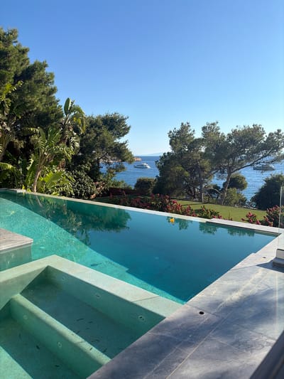 This is a high-angle, eye-level medium shot of an infinity pool overlooking the sea. The pool is filled with turquoise water and has a built-in spa area with steps. The water reflects the clear blue sky and lush green trees. In the background, there is a body of water with several boats, and beyond that, a coastline with more trees and buildings. The overall scene depicts a luxurious vacation spot with a focus on outdoor leisure and scenic views. There are no people visible in the image.