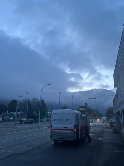A vertical shot captures a cloudy street scene dominated by a large mountain veiled in mist. A white JCDecaux van occupies the foreground, its back doors open, with a person leaning into the vehicle. Street lights punctuate the roadside, and the road markings suggest a wet surface. Buildings and vegetation line the street, contributing to the overcast and somewhat gloomy atmosphere.