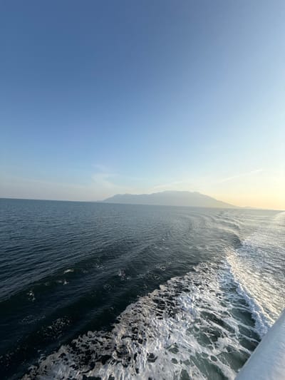 An elevated, wide-angle view captured offshore from Kato Karioutes, Greece, depicts an expanse of open sea and sky under clear conditions. The foreground features dark blue-green water with a distinct white foamy wake originating from the bottom right corner and extending diagonally towards the mid-right. A segment of a white structure, consistent with a vessel's hull or railing, is visible at the bottom right. Undulating water surfaces span the middle ground. In the background, a range of hazy mountains or hills stretches across the horizon line from the left-center to the far right. The sky, occupying the upper portion of the frame, is clear and transitions from a medium blue overhead to a lighter, faintly sunlit blue-yellow hue towards the right horizon. No individuals or specific objects beyond the natural environment and vessel wake are discernible.