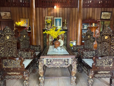 This image captures the interior of a traditionally styled room, likely a living or reception area, in Thới Sơn, Vietnam. The scene is dominated by a grand, elaborately carved wooden furniture set consisting of a large rectangular table with a light-colored marble top and six matching armchairs. The dark wood of the furniture is richly adorned with intricate gold-colored inlay patterns depicting flowers, leaves, and possibly birds. A small white ceramic teapot and teacups are arranged on the table's marble surface.
Against the back wall, which is paneled with dark wood, there is a central altar-like display cabinet. This cabinet holds various items, including multiple vases filled with vibrant yellow flowers, brass candle holders or lamps, and framed portraits. Two prominent framed portraits on the cabinet depict spiritual figures, one with a halo, possibly Guanyin. Another smaller framed portrait is visible inside the display cabinet on the far right. A clock face is partially visible on the lower section of the central display. On either side of the central display, there are ornate wooden cabinets, one of which appears to be a glass-fronted display cabinet on the right, containing various decorative objects and another portrait.
The room features dark wooden columns and a matching wooden ceiling with decorative carved elements. The floor is tiled in a light brown or beige color, simulating wood grain. Framed pictures hang on the walls; one on the left clearly shows text "Việt Nam," depicting a building, and another on the right features a landscape or architectural drawing. Green curtains are visible behind the right-hand cabinet.
There are no people present, and no specific activity is taking place, but the setup, especially the arrangement of flowers and spiritual portraits, suggests a prepared space for reflection, veneration, or receiving guests, possibly for a special occasion like Tết (Vietnamese New Year) given the prominent yellow flowers often associated with this celebration. The room exudes a sense of traditional elegance, reverence, and cultural richness. Visible text includes "Việt Nam" on a picture frame on the left and "VIETNAM" and clock numbers on the central altar.