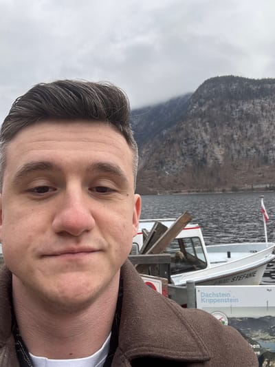 A selfie-style portrait captured outdoors in Hallstatt, Austria, shows a male subject in the foreground. The subject has short, neatly styled dark brown hair, a fair complexion, and is looking directly at the camera. He is wearing a dark brown, possibly wool, collared coat over a white garment, with a black strap visible on his left shoulder.

In the mid-ground, a dark, rippled body of water, Hallstätter See, is visible. Moored on the water is a white motorboat named "STEFANIE" with the registration "O-13.322" prominently displayed. The boat features a small cabin and contains some wooden planks or structures. A small flag, resembling the Austrian flag (red and white), flies from the boat's stern. Partially visible in the lower right, a sign with "Dachstein . Krippenstein" and a mountain graphic further confirms the regional location. A dark wooden pier or dock structure is visible between the man and the boat.

In the far background, wooded mountains rise, their upper slopes partially obscured by low-hanging clouds or mist. The trees on the mountains appear bare or sparse, suggesting a cool season. The sky is overcast with a uniform grey cloud cover.