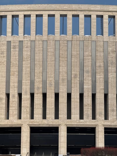 This image displays the facade of a multi-story building located in Plodorodny, Russia. The uppermost visible section features a row of nine tall, narrow, rectangular windows, through which a clear blue sky is visible. White accumulations, consistent with snow, rest on the ledges directly beneath these windows. The central facade consists of light-colored, vertically textured stone, possibly travertine. This section is characterized by a series of evenly spaced vertical elements, each comprising multiple narrow, dark vertical stripes or louvers. Beneath these striped sections are deeper, recessed vertical openings, appearing dark. Between these dominant vertical features, subtle, repeating decorative patterns are embossed or carved into the stone surface, resembling stylized floral or geometric motifs. The lowest visible portion of the building showcases larger, recessed rectangular openings framed by broad, light-colored stone, with dark glass or panels filling these apertures, likely indicating ground-floor entrances or large windows. A small area of reddish-brown vegetation is visible in the bottom right foreground. No individuals are present, and no actions or interactions are depicted within the frame. The scene is illuminated by natural light, suggesting daytime.