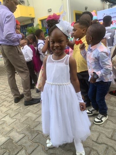 The image shows a group of children and adults standing on a brick pathway. The focus is on a young girl in the center of the image, wearing a white dress with a silver headband, holding her hands to her sides, looking towards the camera. The child is smiling. The girl is standing in front of a group of children, including a boy in a yellow shirt wearing a red bowtie, a boy in a light blue and white checkered shirt with a dark blue bowtie, and other children in white shirts, all in various poses. The girl is standing in front of an adult man wearing a lavender button-down shirt and khaki pants. The background is a blurred out view of buildings and vegetation. The location is in the city of Close, Nigeria, based on the coordinates (latitude: 6.571, longitude: 3.39).