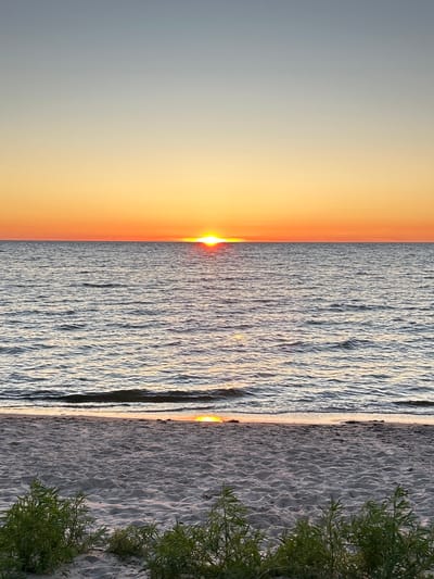 A serene sunset is captured over the Baltic Sea at Meremõisa, Estonia. The sun, a vibrant orange orb, is positioned on the horizon, casting a warm glow across the sky. The sky transitions from a pale blue at the top to a gradient of yellow and orange closer to the horizon. The sea is calm, with gentle ripples reflecting the colors of the sunset. A sandy beach occupies the foreground, with sparse green vegetation growing near the water's edge. The image is a vertical shot, emphasizing the vastness of the sky and sea.