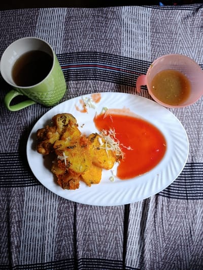 This image depicts a static indoor scene in Vanga ward, Kenya, presenting a meal laid out on a patterned fabric surface. The primary subject is a white oval plate, centrally positioned, holding several pieces of golden-brown, irregularly shaped fried food items, consistent with a type of savory fritter. Adjacent to the fried items on the plate is a substantial pool of bright red sauce. A small quantity of shredded white vegetable, possibly cabbage, is scattered near the fried food and the red sauce. To the upper left of the plate, a light green ceramic mug with a handle contains a dark liquid. To the upper right, a light pink plastic cup with a handle contains a light brown, translucent liquid with visible particulate matter. The fabric surface beneath these items displays a repetitive zigzag or chevron pattern in shades of brown, gray, and white, interrupted by horizontal black stripes with thin red and blue lines. No individuals are visible in the scene.