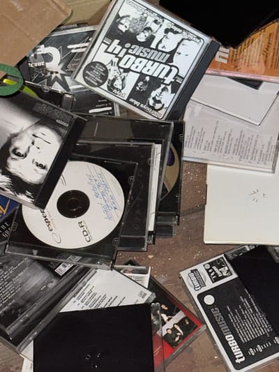 The image displays a chaotic pile of CDs and CD cases scattered on a rough, dusty surface that appears to be a floor or a cluttered tabletop. The items are various, including what appear to be music albums, blank CD-Rs with handwritten labels, and cases for these discs. One CD case features a black and white image of four individuals and the text "Turbo Music". Another CD-R has a handwritten label that seems to include "Metallica", "300", and "Mylene Farmer". In the background, a cardboard box and what looks like a piece of black plastic or electronics are visible. The lighting is uneven, suggesting an indoor environment with ambient light. There are no people visible in the image. The scene appears to be a collection of old or discarded media. The reference to Chernyakhovsk, Russia, is not visually supported by any elements within the provided image.