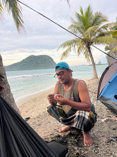 A man wearing a teal baseball cap and glasses crouches on a pebble beach, holding a colorful, striped sphere. He is dressed in a grey tank top and a patterned sarong. To his left is a black hammock and a portion of a palm tree. To his right, a blue and grey tent is set up. In the background, turquoise waves crash onto the shore, and a large, verdant hill rises from the sea under a cloudy sky. The scene is set in Ujongsedheun, Indonesia.
