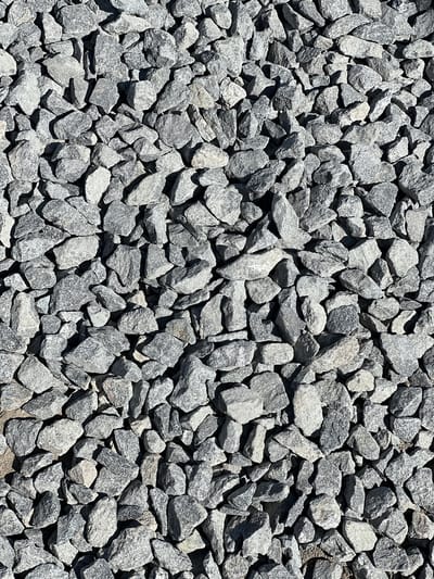 The image is a close-up, overhead view of a surface covered with a dense layer of gray, irregularly shaped crushed stone. The stones vary in size, with some appearing larger and more angular than others. The texture is rough and uneven, with shadows cast by the stones indicating a light source from above and slightly to the side. The overall impression is that of a construction or landscaping material. There is no indication of any specific location in Újezd u Brna, Czechia, nor are there any people, actions, or interactions depicted.
