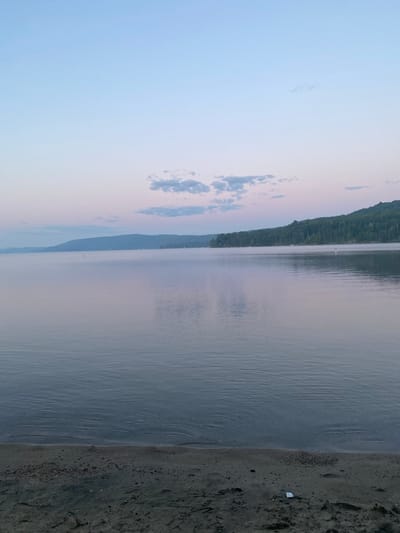 An atmospheric long-shot photograph captures a serene lake at dawn, with a light mist hovering over the water. In the distance, a tree-lined shore with rolling hills forms the backdrop. The sky transitions from a pale blue at the top to soft pink and lavender hues near the horizon, with scattered wispy clouds. In the foreground, a dark, sandy beach is visible, with small pebbles and sand grains. A white object, possibly a wrapper, lies on the sand near the water's edge. The scene suggests a tranquil natural environment, likely in Duhamel, Canada, given the presence of a lake and lush vegetation.