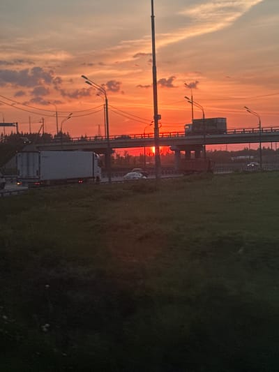 The image shows a sunset in посёлок опытного хозяйства «Ермолино», Russia. A highway overpass with streetlights is visible. There is a truck on the road below and a car on the highway above. The sky is filled with orange and yellow clouds. The grass is green.