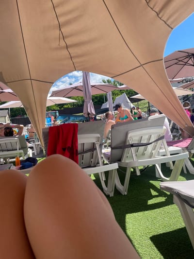The image depicts an outdoor scene at a swimming pool complex in Zazymia, Ukraine. The foreground is dominated by the legs of a person, positioned in a way that suggests they are reclining on a lounge chair.
In the mid-ground, several white plastic lounge chairs are visible, some occupied by people. A red towel with a dark heart design hangs on the back of one chair. Further back, there are more lounge chairs and umbrellas providing shade. A large, light-colored canopy stretches across the top of the image, creating a shaded area.
In the background, a swimming pool is partially visible with inflatable toys, suggesting recreational activities. The surrounding environment includes trees and buildings, indicating a developed outdoor space. The sky is blue with some clouds. People are seen in the pool area and on lounge chairs, some interacting with each other. The overall atmosphere is that of a sunny day at a leisure facility.