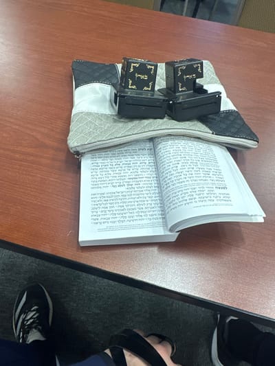 On a reddish-brown wooden table in Medford, United States, a pair of black tefillin are positioned on a multi-paneled pouch, colored black, white, and a light textured gray/beige. The pouch is partially unzipped and rests on an open white prayer book, displaying Hebrew text across both visible pages. The book's spine is oriented towards the viewer. Below the table, a dark gray carpeted floor is visible. Two feet, likely belonging to the same individual, are partially visible. The left foot wears a black and white athletic shoe, while the right foot wears a dark-colored sandal. No other individuals or actions are depicted.