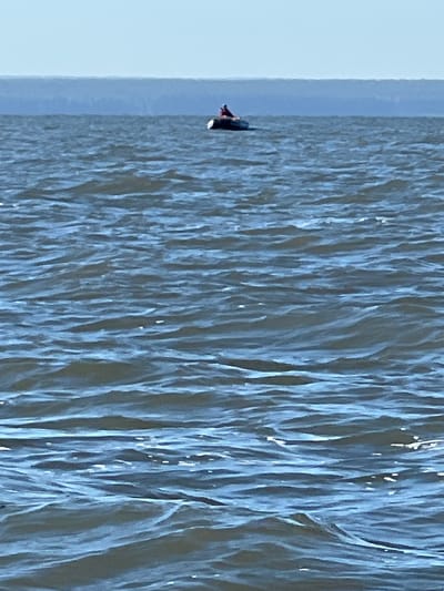 A person is sitting in a small boat on a body of water. The boat is dark in color and the person is wearing red clothing. The water is choppy with small waves. In the background, there is a hazy horizon line, suggesting land or a larger body of water. The sky is clear and blue. The scene appears to be set outdoors in a natural environment.