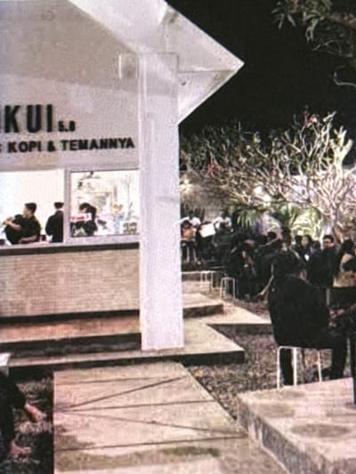 The image depicts an outdoor nighttime scene at a cafe called "KUI..KOPI & TEMANNYA" in Babahjurong, Indonesia. A portion of the cafe's facade is visible, featuring a sign with the establishment's name. Several people are gathered, with some seated and others standing. In the foreground, there appears to be a low wall or counter with a few individuals. Further back, a larger group of people are congregating under trees, suggesting an outdoor seating area. The lighting is dim, characteristic of a nighttime setting, with some artificial lights illuminating the scene. The overall atmosphere appears to be social, with patrons enjoying the cafe's ambiance.