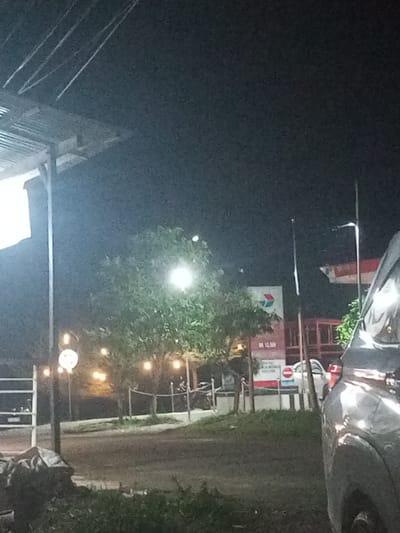 The image depicts a night scene in Lawang, Indonesia, possibly at or near a gas station. The composition is poorly lit. A silver vehicle is situated in the foreground on the right side. A gas station sign and building can be seen in the middle ground. Illumination is present from streetlights and strings of lights strung through trees. Fencing is visible. Patches of foliage appear along the lower edge. The time of day is night.