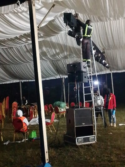 The scene depicts an outdoor event setup at night under a large white marquee tent with a draped fabric ceiling, situated on a grassy ground in Marenyo-Shianda ward, Kenya. Artificial light sources illuminate the interior.
In the central foreground, a male individual, wearing a dark jumpsuit with a reflective yellow safety vest bearing the word "Spitfire" on the back, stands on the upper steps of a silver A-frame ladder. He is actively adjusting a black lighting fixture and an associated black speaker unit, both attached to a metal truss structure suspended from the tent's internal framework. Below the ladder, a stack of three black audio speakers is positioned, with a larger black speaker cabinet resting directly on the ground beside the ladder's base.
Two additional male individuals are present near the bottom of the ladder. One wears a shiny silver jacket, dark pants, and a dark cap. The other wears a red jacket, blue jeans, and holds a light blue item, possibly a bottle or small bag, in his left hand.
To the left of the main setup, a person wearing a white head covering and an orange reflective safety vest is seated on a red chair with a gold frame, facing away from the camera. A green bag rests on the ground beside this person. Further in the background, to the left, a fourth individual is partially visible behind a table laden with electronic equipment, possibly a sound mixer.
Numerous empty red chairs with gold frames are scattered or stacked throughout the grassy area, suggesting preparations or post-event activities. A large green sack or bag is also visible on the grass in the mid-ground. Various cables are evident, connected to the audio and lighting equipment.