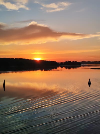 This is a photograph of a serene body of water during a sunset in Терехово, Russia. The sun is low on the horizon, casting a warm orange glow across the sky and reflecting on the water's surface. Silhouetted trees line the distant shore, creating a dark, undulating border against the vibrant sky. Two buoys are visible in the water; one is a dark, conical marker on the left, and the other is a lighter, cone-shaped marker with a light on top, positioned further to the right. The foreground of the water shows the distinct pattern of ripples, suggesting movement from a boat or the wind. The overall scene is tranquil and bathed in the soft light of twilight.
