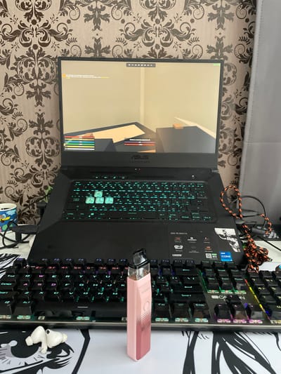 The image shows a gaming laptop on a desk, displaying a game with a user interface. The laptop is open, with the screen visible and the keyboard illuminated by RGB lighting. To the right of the laptop, there is a charging cable with an orange and black pattern. In front of the laptop, there is a pink vape device and a pair of white wireless earbuds. The desk is covered with a patterned mousepad featuring black and white graphics. The background consists of a wallpaper with a damask pattern. The scene suggests a personal gaming setup. There is no indication of the specific location within Азершчынскі сельскі Савет, Belarus.